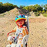 bike_seat, blue_sky, child, colorful, daylight, dirt_path, happy, helmet, nature, outdoor, overalls, person, playful, safety_harness, seat, smiling, sunny, toddler, trees, young_child