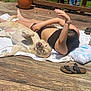dog, person, sunbathing, deck, towel, pillow, sandals, potted_plant, grass, wooden_floor, relaxation, sunny, outdoor, pet, companion, leisure, summer, tongue_out, casual, barefoot