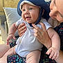 baby, child, hat, blue_hat, onesie, smiling, adult, hands, outdoor, cushion, chair, stone_wall, person, toddler, happy, skin, face, holding, sunlight, casual_clothing