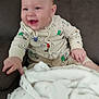baby, child, smiling, laughing, onesie, dog_pattern, blanket, couch, furniture, indoor, happy, cute, infant, person, soft_texture, hand, face, head, seated, cozy
