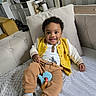 Soan a rejoint le concours — aidez-le/la à gagner de superbes lots ! baby, blanket, brown_pants, casual_clothing, child, couch, cozy, cute, footwear, happy, home, indoor, living_room, person, pillow, relaxed, smile, toddler, toy, yellow_jacket