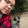 person, dog, selfie, outdoor, sidewalk, garden, trees, glasses, plaid_shirt, blue_heeler, australian_cattle_dog, black_white_coat, sitting, leash, portrait, close_up, pet, companion, daylight, humorous