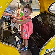 Alliyah Johnson is registered to the contest to win money with this photo: toddler, child, pink_dress, yellow_car, kiddie_ride, indoor, steering_wheel, adult, person, footwear, curly_hair, serious_expression, play_area, floor, metal_plate, poster, shopping_mall, toy_car, hand, casual_clothing