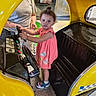 toddler, child, pink_dress, yellow_car, kiddie_ride, indoor, steering_wheel, adult, person, footwear, curly_hair, serious_expression, play_area, floor, metal_plate, poster, shopping_mall, toy_car, hand, casual_clothing