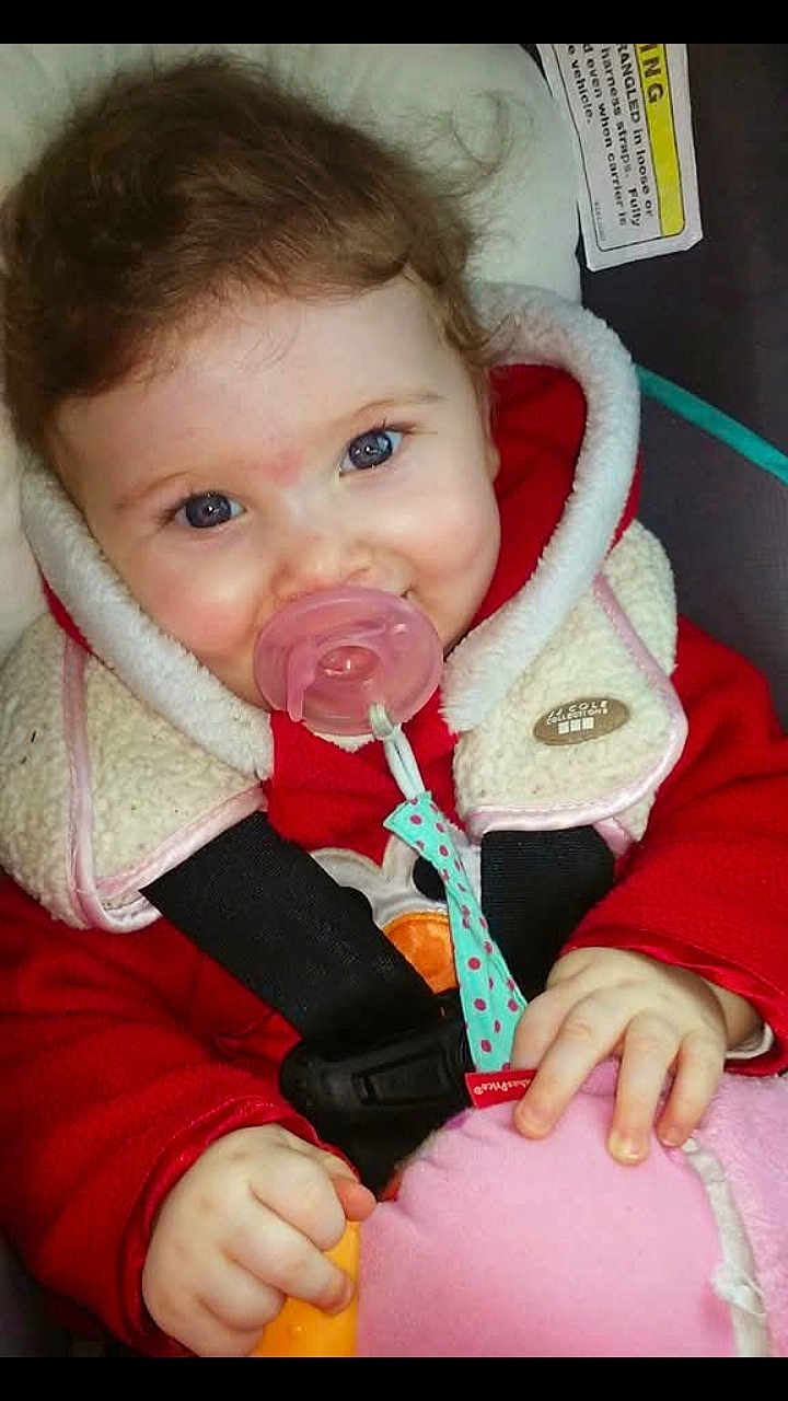 Karli Mae joined the competition — help win amazing prizes! baby, child, pacifier, red_clothing, fleece_collar, curly_hair, blue_eyes, car_seat, toy, hand, cute, infant, smiling, cozy, indoor, person, closeup, face, warm_clothing, seatbelt