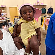 Yeshua is registered to the contest to win money with this photo: baby, child, person, clothing_store, yellow_onesie, pink_cap, adult_hand, jewelry, socks, shopping, rack, clothes, retail, indoor, curious, expression, casual, portrait, holding, fashion