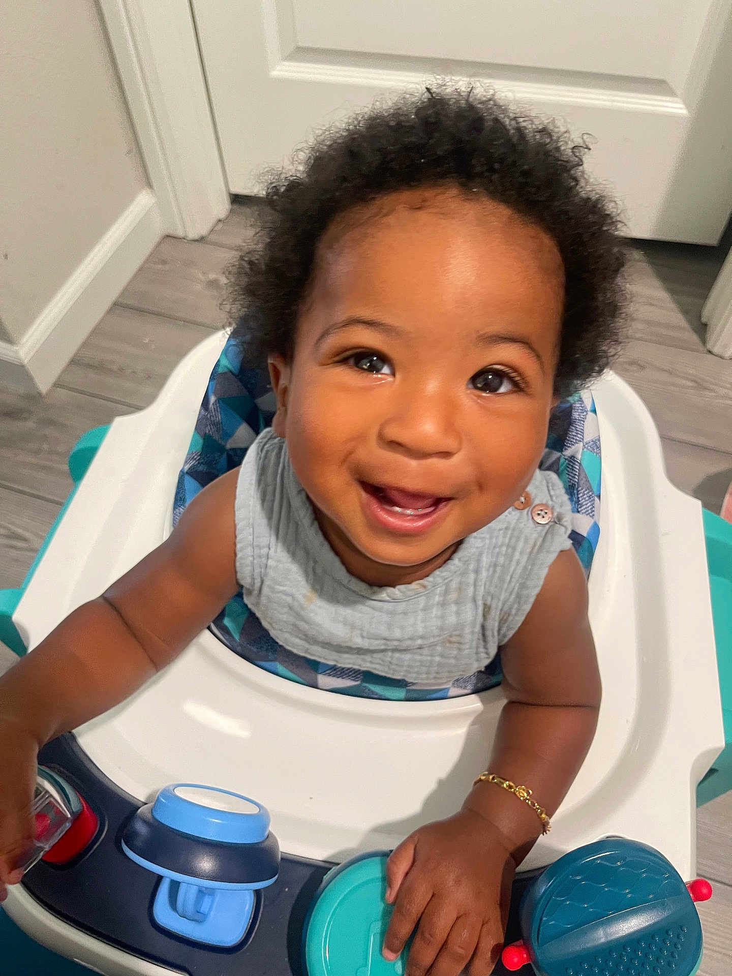 Suede is registered to the contest to win money with this photo: toddler, child, smiling, high_chair, toy, indoor, happy, face, curly_hair, arm, clothing, floor, door, bracelet, baby, play, seat, person, expression, closeup