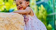 Neriah is registered to the contest to win money with this photo: toddler, child, smiling, curly_hair, rock, outdoor, sunlight, greenery, trees, building, casual_clothing, white_shoes, playful, happy, summer, daylight, cute, portrait, nature, urban