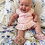 baby, smiling, pink_dress, shoes, floral_bedspread, infant, cute, happy, sitting, indoors, child, portrait, face, head, legs, arms, clothing, footwear, bedroom, home