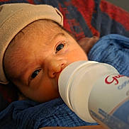 Raphaël a rejoint le concours — aidez-le/la à gagner de superbes lots ! baby, newborn, infant, feeding_bottle, bottle, hat, swaddle, blanket, close_up, portrait, face, eyes, hand, mouth, skin, lying_down, cozy, indoors, text_on_bottle, background_fabric