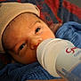baby, newborn, infant, feeding_bottle, bottle, hat, swaddle, blanket, close_up, portrait, face, eyes, hand, mouth, skin, lying_down, cozy, indoors, text_on_bottle, background_fabric
