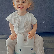 Syanna a rejoint le concours — aidez-le/la à gagner de superbes lots ! toddler, child, smiling, indoor, potty_chair, bunny_shape, white_shirt, pants, sandals, hair_bun, floor, couch, happy, cute, childhood, playful, toys, home, furniture, portrait