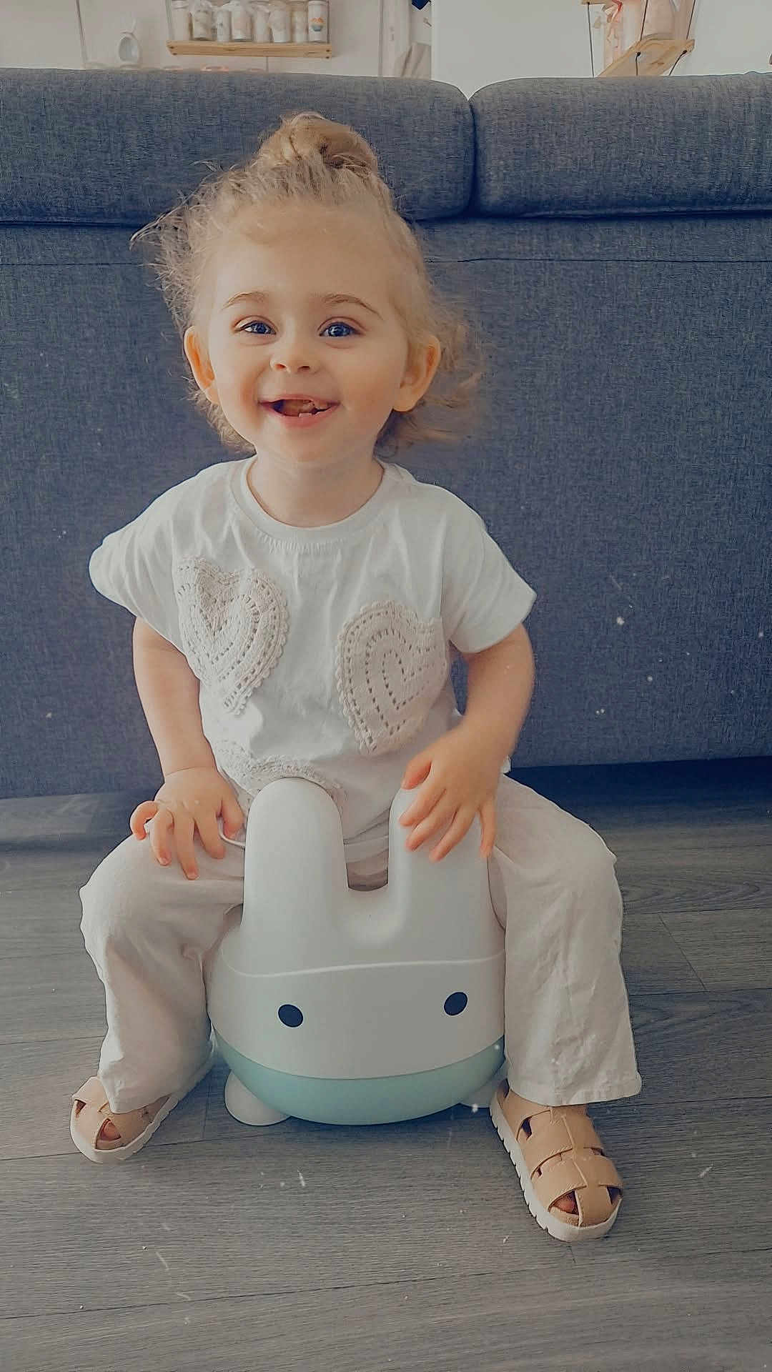 Syanna a rejoint le concours — aidez-le/la à gagner de superbes lots ! toddler, child, smiling, indoor, potty_chair, bunny_shape, white_shirt, pants, sandals, hair_bun, floor, couch, happy, cute, childhood, playful, toys, home, furniture, portrait