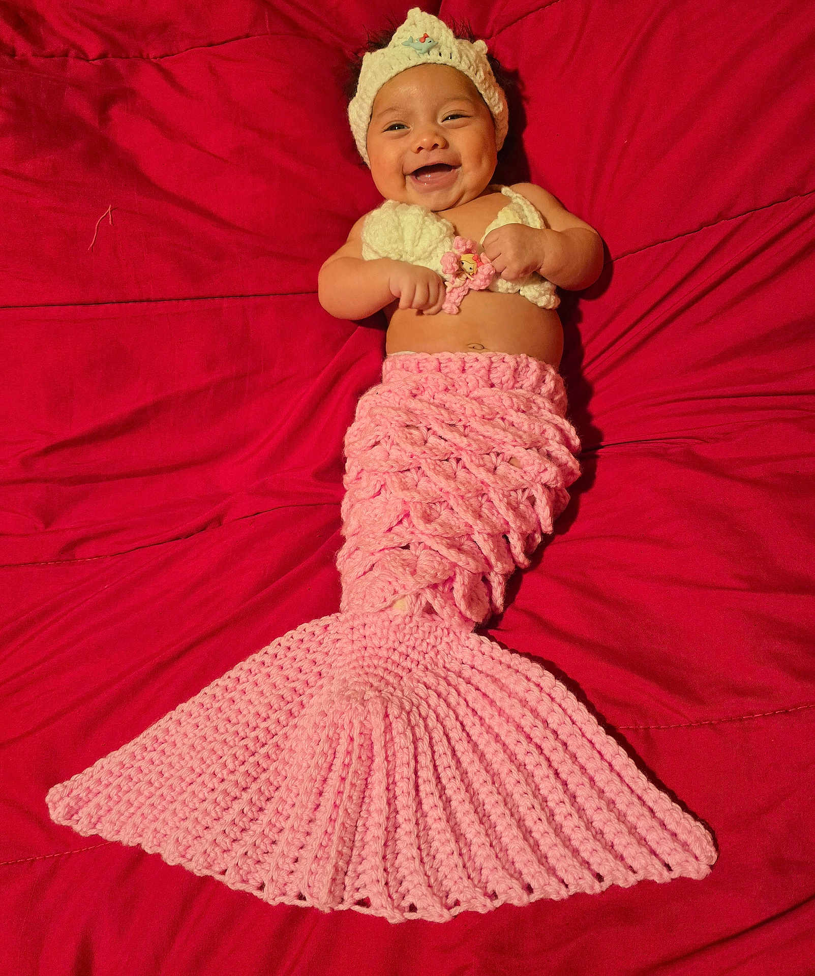Andrea is registered to the contest to win money with this photo: baby, smiling, crocheted, mermaid, pink, headband, blanket, red, cute, infant, happy, costume, handmade, portrait, lying_down, child, toddler, knitwear, cozy, playful