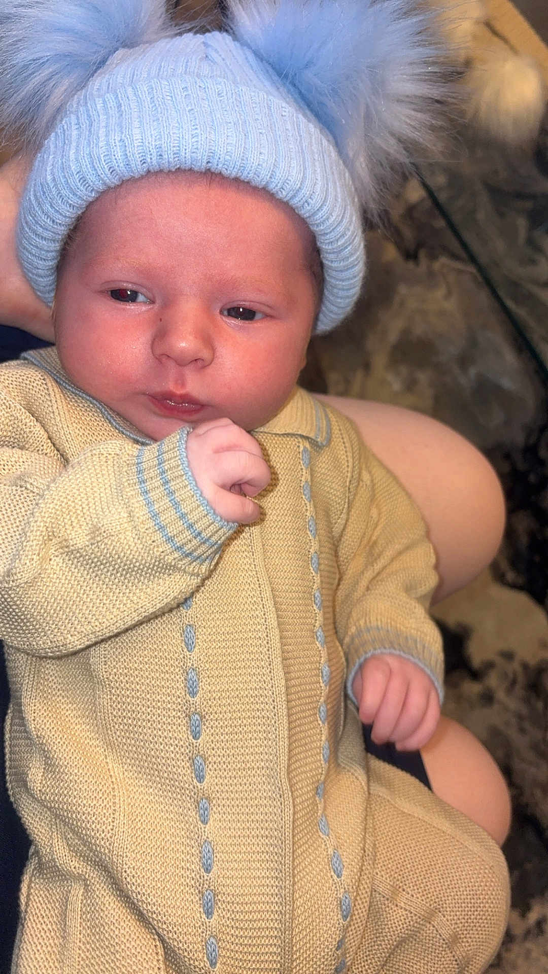 Tommy joined the competition — help win amazing prizes! baby, infant, knitwear, hat, pom_pom, yellow_clothing, blue_hat, hand, face, person, sitting, close_up, cute, indoor, soft_texture, warm_clothing, portrait, newborn, skin, curious