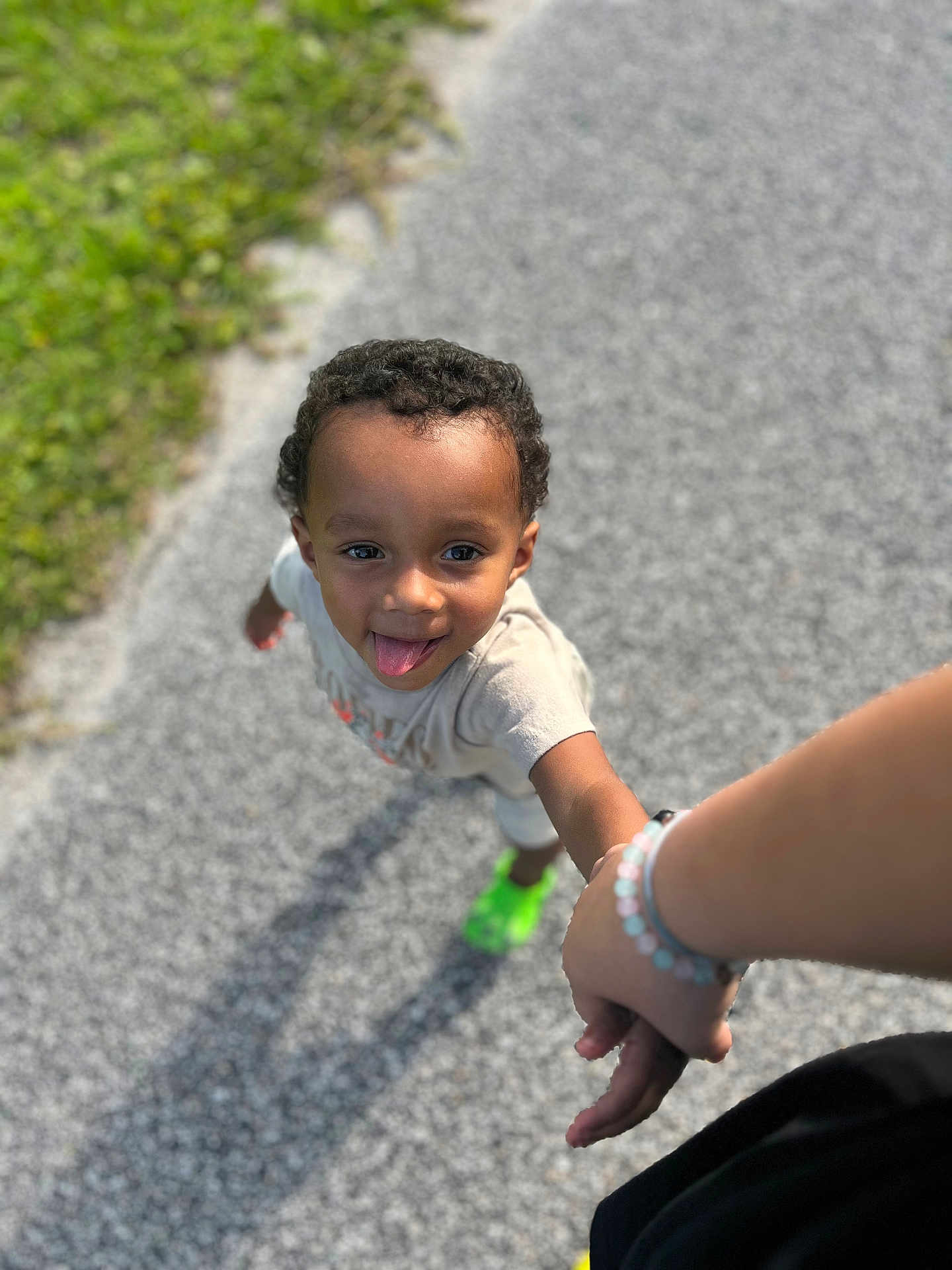 Marcus is registered to the contest to win money with this photo: child, toddler, playing, holding_hand, curly_hair, outdoor, pathway, gravel, greenery, sunlight, tongue_out, happy, casual_clothing, sneakers, person, walking, joyful, shadow, bracelet, summer