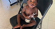 Zarie joined the competition — help win amazing prizes! toddler, child, diaper, chair, folding_chair, table, floor, indoor, baby, food, messy, utensil, black_chair, curious, expression, person, skin, hair, looking_up, cute