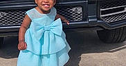 Mariah is registered to the contest to win money with this photo: toddler, child, blue_dress, bow, smiling, standing, outdoor, car, luxury_car, black_car, concrete, sunlight, shoes, happy, cute, fashion, young_child, person, vehicle, portrait