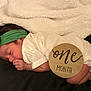 baby, sleeping, infant, headband, white_clothing, blanket, wooden_sign, one_month, newborn, cute, peaceful, indoors, portrait, child, resting, hand, face, soft_texture, black_surface, milestone