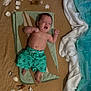 baby, child, swim_shorts, towel, cardboard, shark_fin, seashells, sand, fabric, blue, white, lying_down, indoors, playful, cute, infant, summer_theme, bare_chest, relaxed, decorative