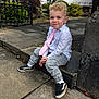 child, boy, curly_hair, white_shirt, pink_tie, grey_pants, black_shoes, outdoor, stone_step, sidewalk, gate, greenery, building, casual, formal, portrait, smile, young, urban, daylight