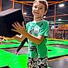 child, boy, smiling, indoor, trampoline_park, playground, foam_block, jumping, recreation, green_mats, orange_rails, sports_facility, short_hair, green_shirt, patterned_shorts, portrait, standing, playful, background_people, safe_play