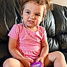 child, toddler, portrait, curly_hair, smiling, serious_expression, pink_clothing, floral_pattern, purple_toy, sitting, couch, black_leather, indoor, home, close_up, face, arms, legs, playtime, photography