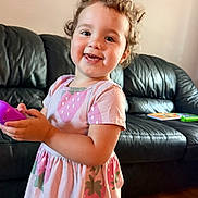Emilia And Olivia is registered to the contest to win money with this photo: child, toddler, girl, smiling, portrait, curly_hair, pink_dress, strawberry_pattern, toy, purple_toy, sitting, standing, couch, leather_sofa, living_room, indoor, wooden_floor, happy, baby_teeth, family_photo