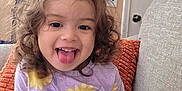 Kilanna joined the competition — help win amazing prizes! casual, child, couch, curly_hair, cute, furniture, happy, home, indoor, orange_pillow, person, playful, portrait, purple_clothing, sitting, smiling, toddler, tongue_out, yellow_flowers, young_child