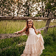 Mirabella is registered to the contest to win money with this photo: child, girl, dress, flower_crown, fence, grass, field, trees, sunlight, outdoor, nature, portrait, smile, long_hair, greenery, sunset, rural, happy, summer, scenic