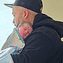 newborn, baby, man, person, black_hoodie, black_cap, hospital_blanket, sleeping, face, head, side_profile, indoors, light, window_light, tender_moment, father_and_child, close_up, human, skin, portrait