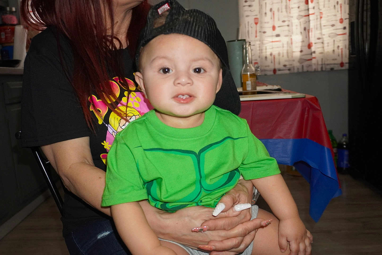 Mkilye joined the competition — help win amazing prizes! toddler, child, person, indoor, cap, green_shirt, adult, hand, table, curtain, floor, chair, nail_art, casual_clothing, face, smile, sitting, background, bottle, kitchen
