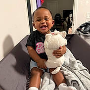 Bella joined the competition — help win amazing prizes! toddler, child, smile, stuffed_animal, toy, sitting, blanket, cushion, socks, indoor, happy, person, clothing, shorts, black_shirt, home, living_room, furniture, face, cute