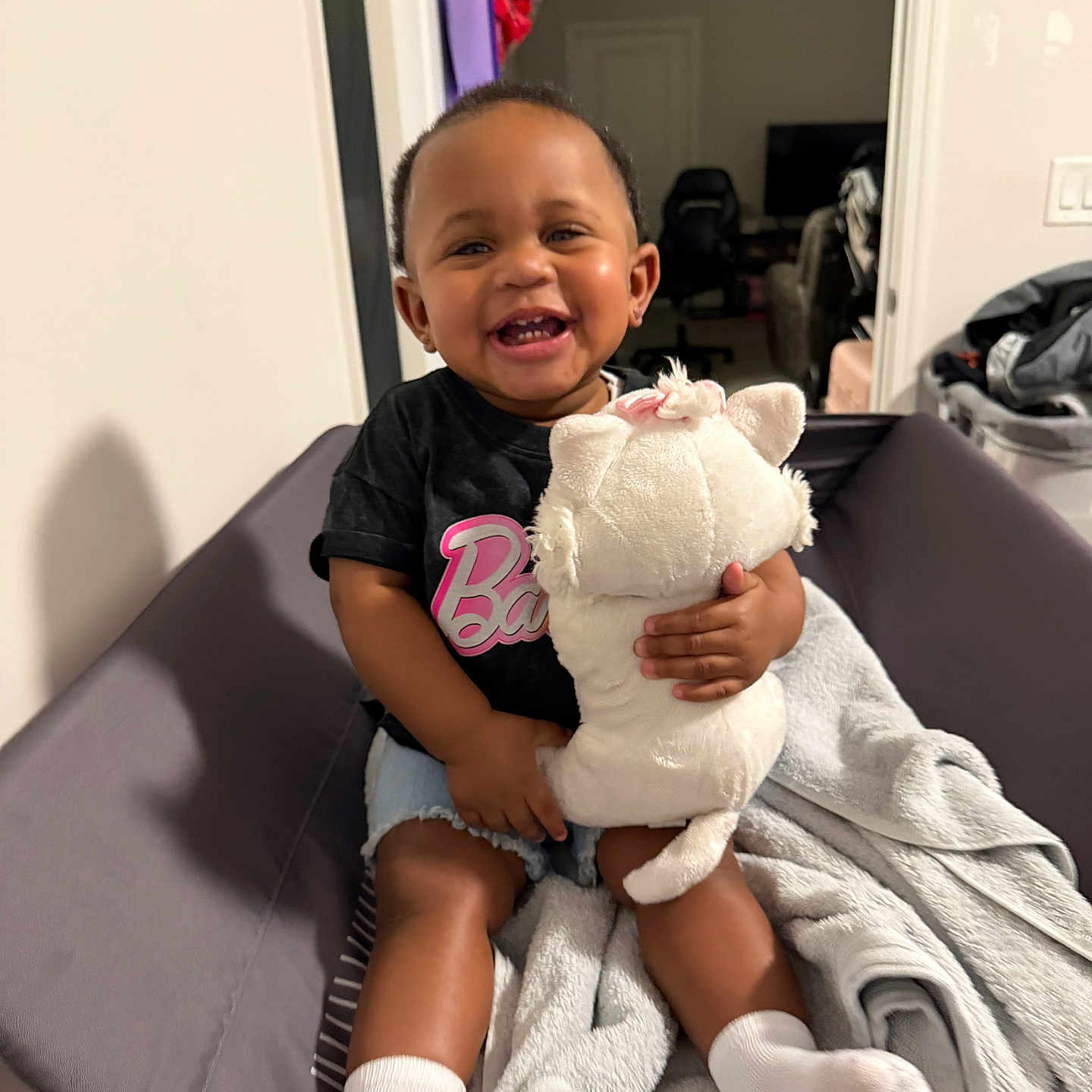 Bella joined the competition — help win amazing prizes! toddler, child, smile, stuffed_animal, toy, sitting, blanket, cushion, socks, indoor, happy, person, clothing, shorts, black_shirt, home, living_room, furniture, face, cute