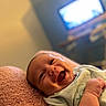 baby, smiling, infant, blanket, pink, lying_down, happy, face, indoors, television, blurred_background, toddler, cute, child, soft_texture, clothing, portrait, skin, hand, expression