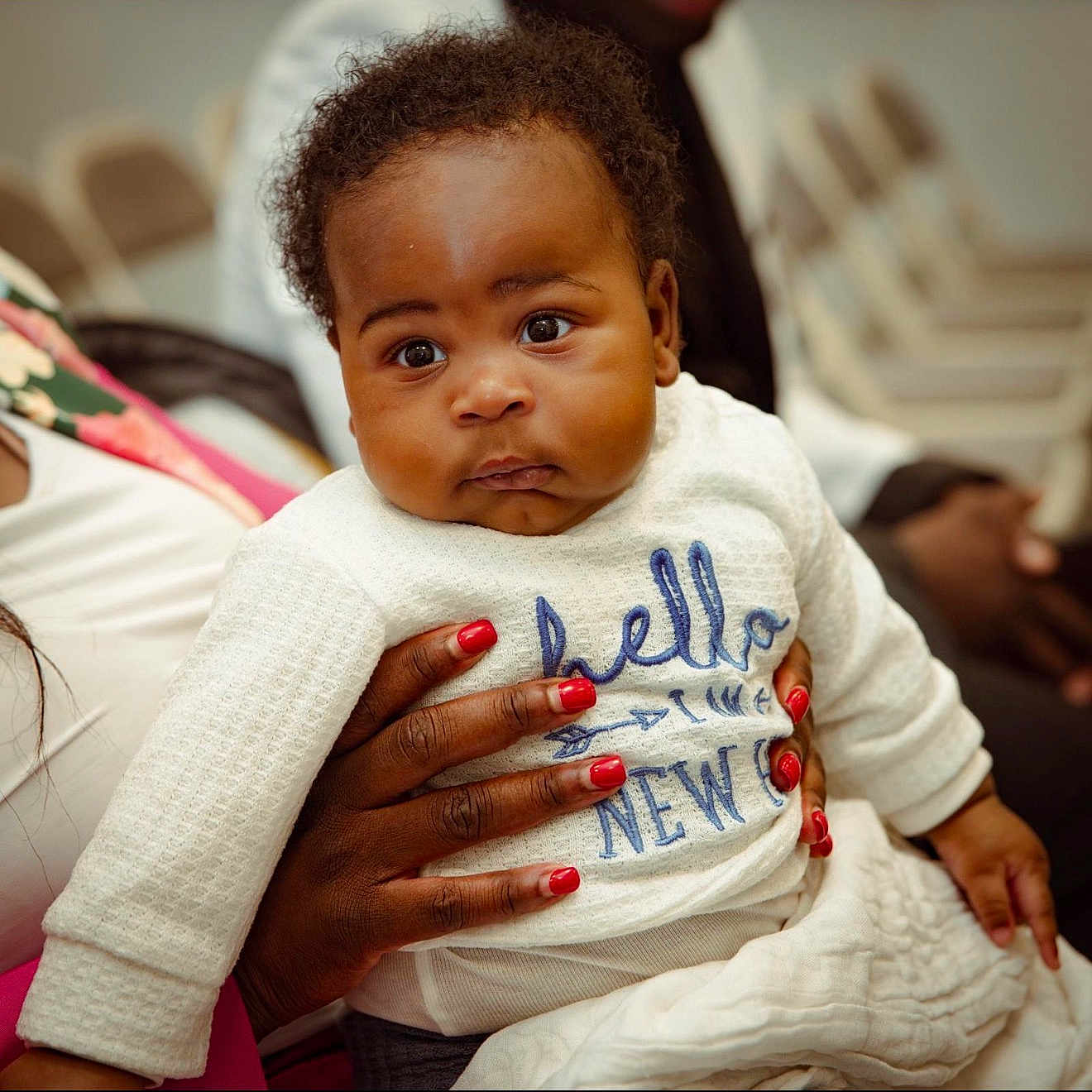 Munyasha Eli joined the competition — help win amazing prizes! baby, infant, child, person, hand, red_nail_polish, curly_hair, white_clothing, embroidered_text, closeup, indoor, seated, skin, face, eyes, expression, holding, caring, warmth, soft_light