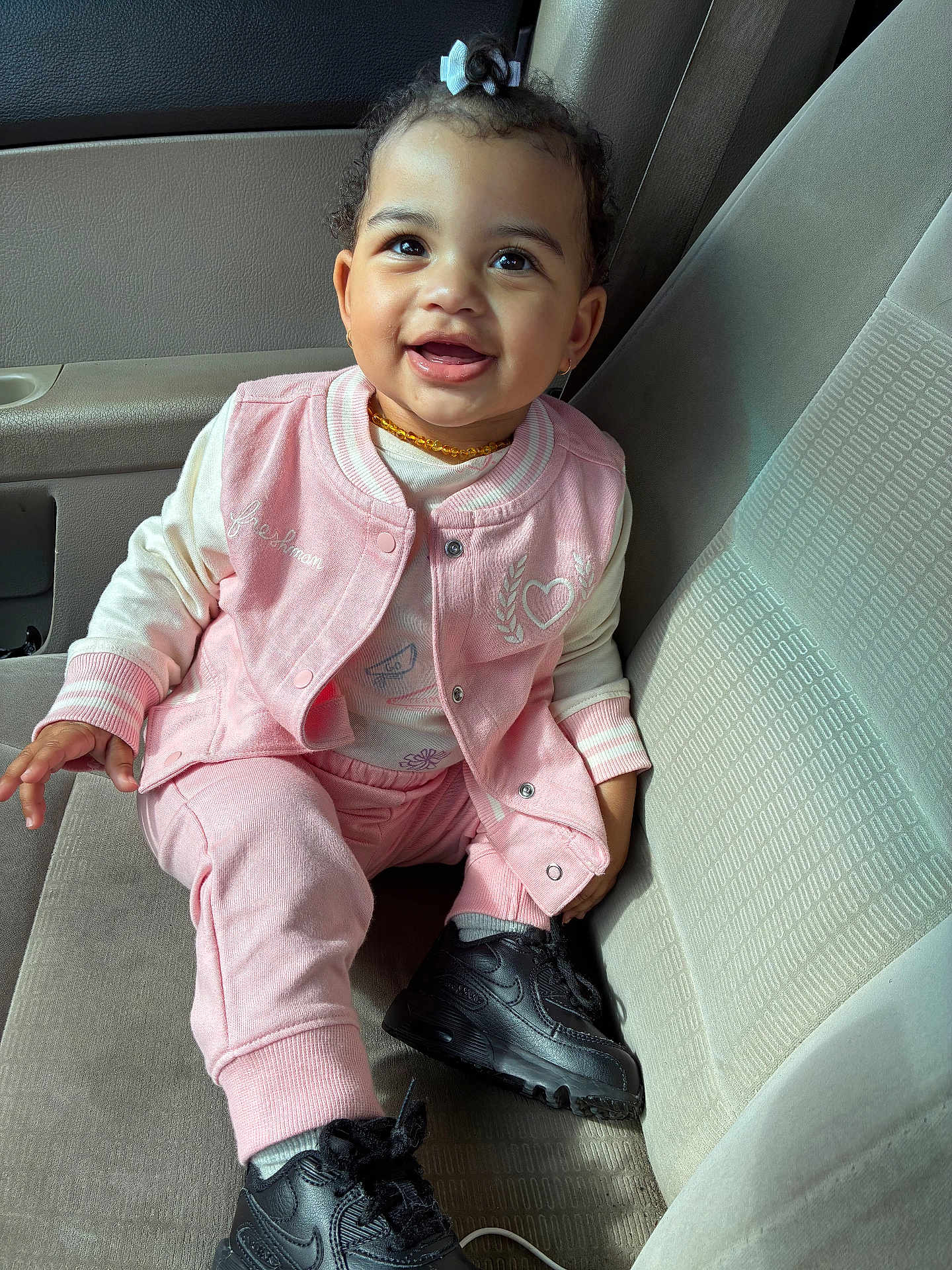 Kaylani joined the competition — help win amazing prizes! toddler, child, smiling, sitting, car_seat, pink_clothing, black_shoes, curly_hair, hair_bow, jacket, pants, happy, person, casual_wear, interior, car, seat, footwear, portrait, cute