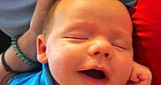 Silas Jericho Day is registered to the contest to win money with this photo: baby, smiling, blue_shirt, hand, bracelet, nails, face, infant, sleeping, closeup, person, skin, fingers, happy, cozy, fabric, background, red, portrait, resting