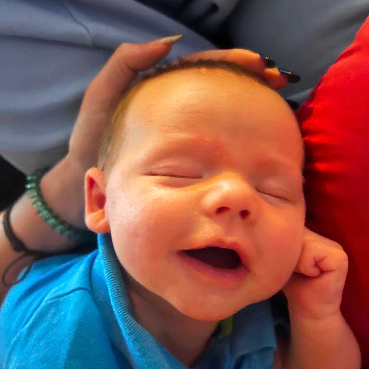 Silas Jericho Day is registered to the contest to win money with this photo: baby, smiling, blue_shirt, hand, bracelet, nails, face, infant, sleeping, closeup, person, skin, fingers, happy, cozy, fabric, background, red, portrait, resting