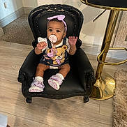 Sevynn is registered to the contest to win money with this photo: baby, child, pacifier, headband, denim_overalls, teddy_bear, chair, black_chair, indoor, wooden_floor, bar_stool, gold, rug, cute, sitting, curious, footwear, pink_shoes, person, infant