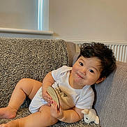 Caleb is registered to the contest to win money with this photo: baby, infant, child, couch, sofa, plush_toy, milestone_plaque, white_onesie, smile, curly_hair, indoors, cozy, texture, soft_light, relaxing, cute, chubby, happy, portrait, home