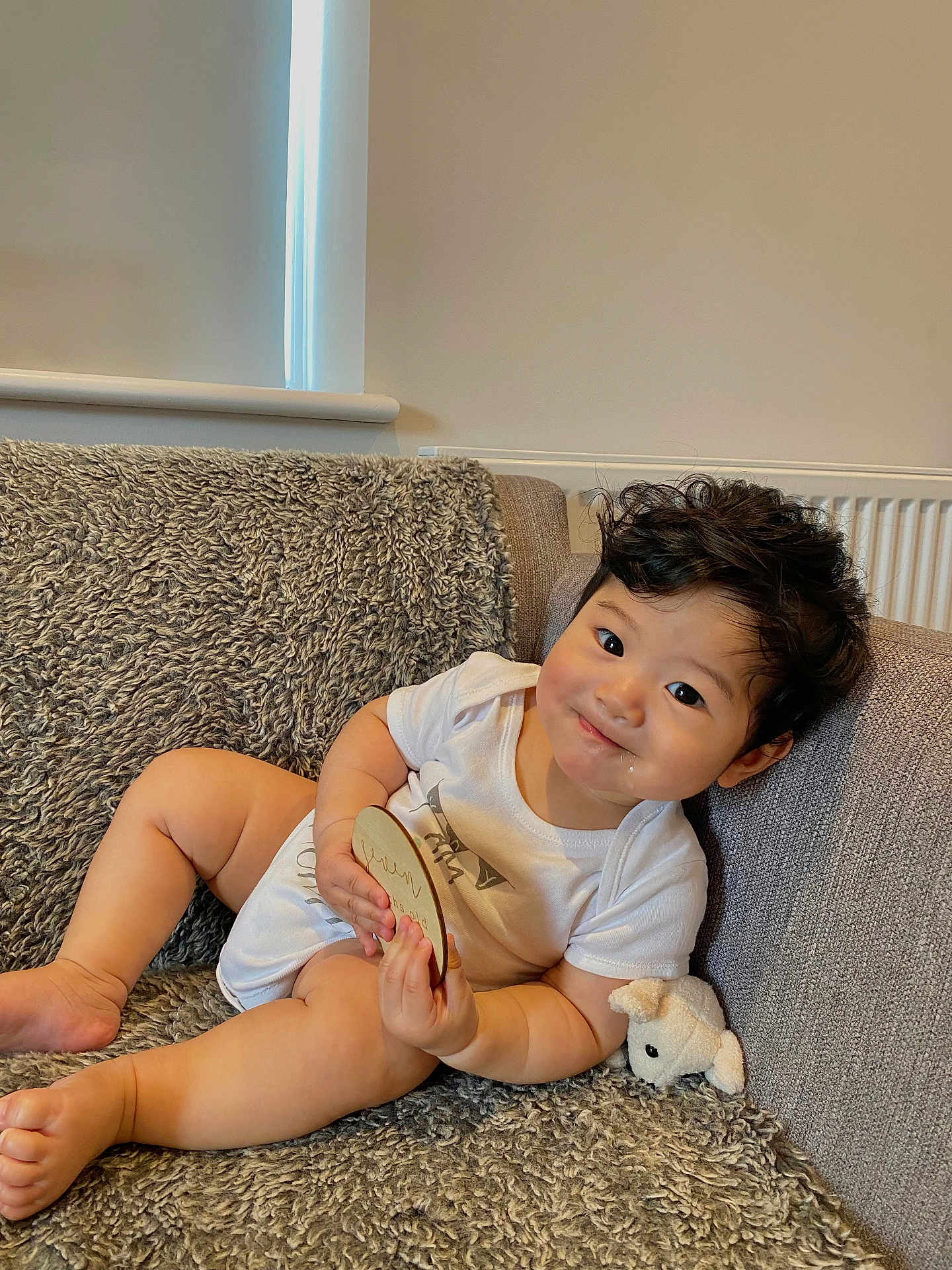 Caleb is registered to the contest to win money with this photo: baby, infant, child, couch, sofa, plush_toy, milestone_plaque, white_onesie, smile, curly_hair, indoors, cozy, texture, soft_light, relaxing, cute, chubby, happy, portrait, home