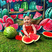 Sophia is registered to the contest to win money with this photo: baby, child, watermelon, fruit, grass, outdoor, smiling, red_clothing, headband, green_bow, food, summer, cute, playful, background, colorful, sitting, nature, happy, portrait