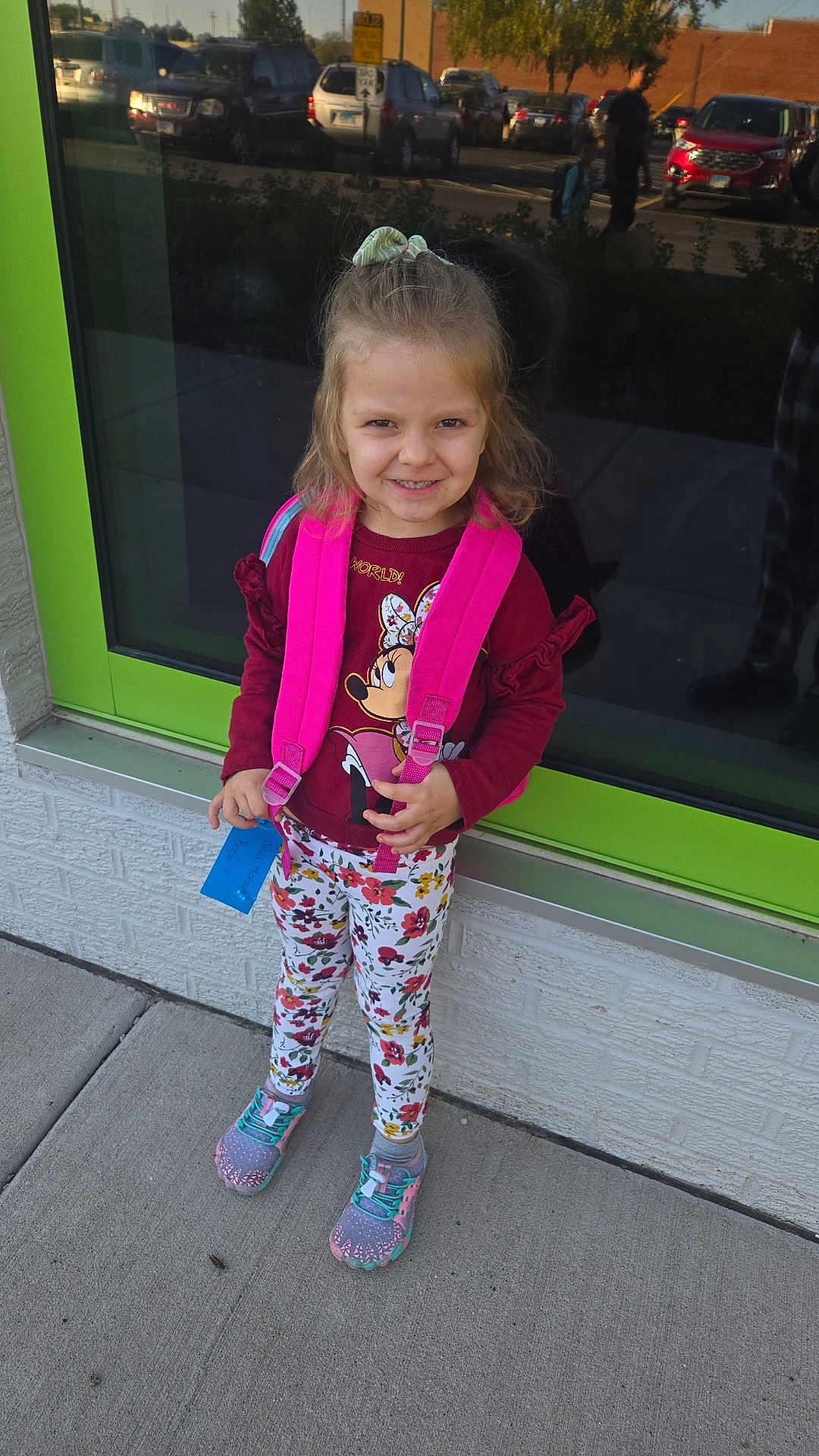 Nova is registered to the contest to win money with this photo: child, girl, backpack, smile, sidewalk, window, reflection, floral_leggings, sneakers, casual_clothing, outdoor, daylight, happy, cute, standing, concrete, urban, playful, young, portrait