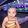 baby, bench, blue_bow, blurred_background, child, colorful_lights, cute, denim_shorts, face, fashion, happy, headband, night, orange_top, outdoor, person, portrait, sitting, smiling, young_child