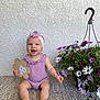 baby, child, smiling, purple_clothing, headband, flower_sign, indoor, sitting, barefoot, happy, pot_of_flowers, decor, texture_wall, rug, cute, infant, portrait, playful, bright, young_child