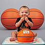 baby, cake, birthday, basketball, celebration, smiling, orange, frosting, hands, face, sitting, toy, happy, child, foot, black_shirt, bracelet, table, indoor, photography