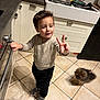 black_pants, boy, cabinet, child, dish_rack, dog, fluffy_dog, gray_shoes, indoor, kitchen, kitchen_counter, oven, peace_sign, sink, small_dog, smiling, standing, sweater, tile_floor, utensils