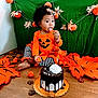 toddler, child, pumpkin_outfit, orange_clothing, striped_pants, cake, chocolate_cake, whipped_cream, autumn_decor, pumpkin_decorations, fall_leaves, wooden_floor, green_backdrop, holiday, festive, cute, playful, sitting, indoor, celebration