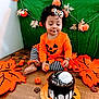 toddler, child, pumpkin_outfit, orange_clothing, striped_pants, fall_decorations, pumpkins, large_leaves, chocolate_cake, whipped_cream, green_backdrop, wooden_surface, holiday_theme, smiling_child, curly_hair, headband, festive, indoors, celebration, cute